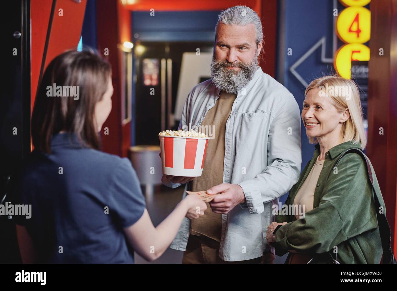 Modern mature man and woman in love passing their tickets for movie to young female cinema hall worker Stock Photo