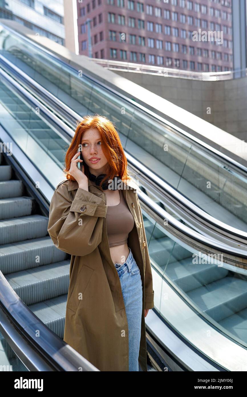 Teen redhead girl talking on cell standing on escalator. Stock Photo