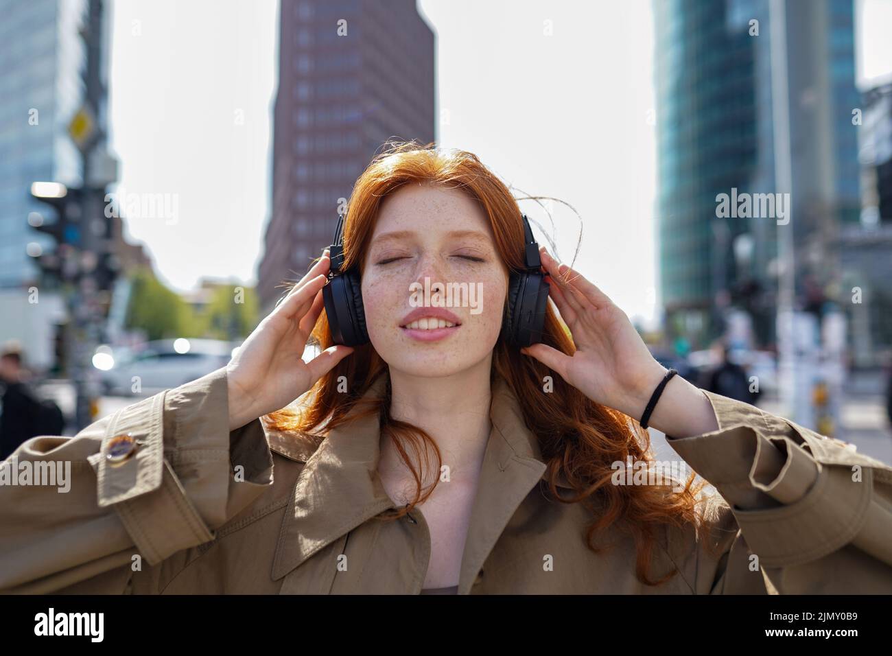 Happy teen redhead girl wearing headphones listening music in big city. Stock Photo