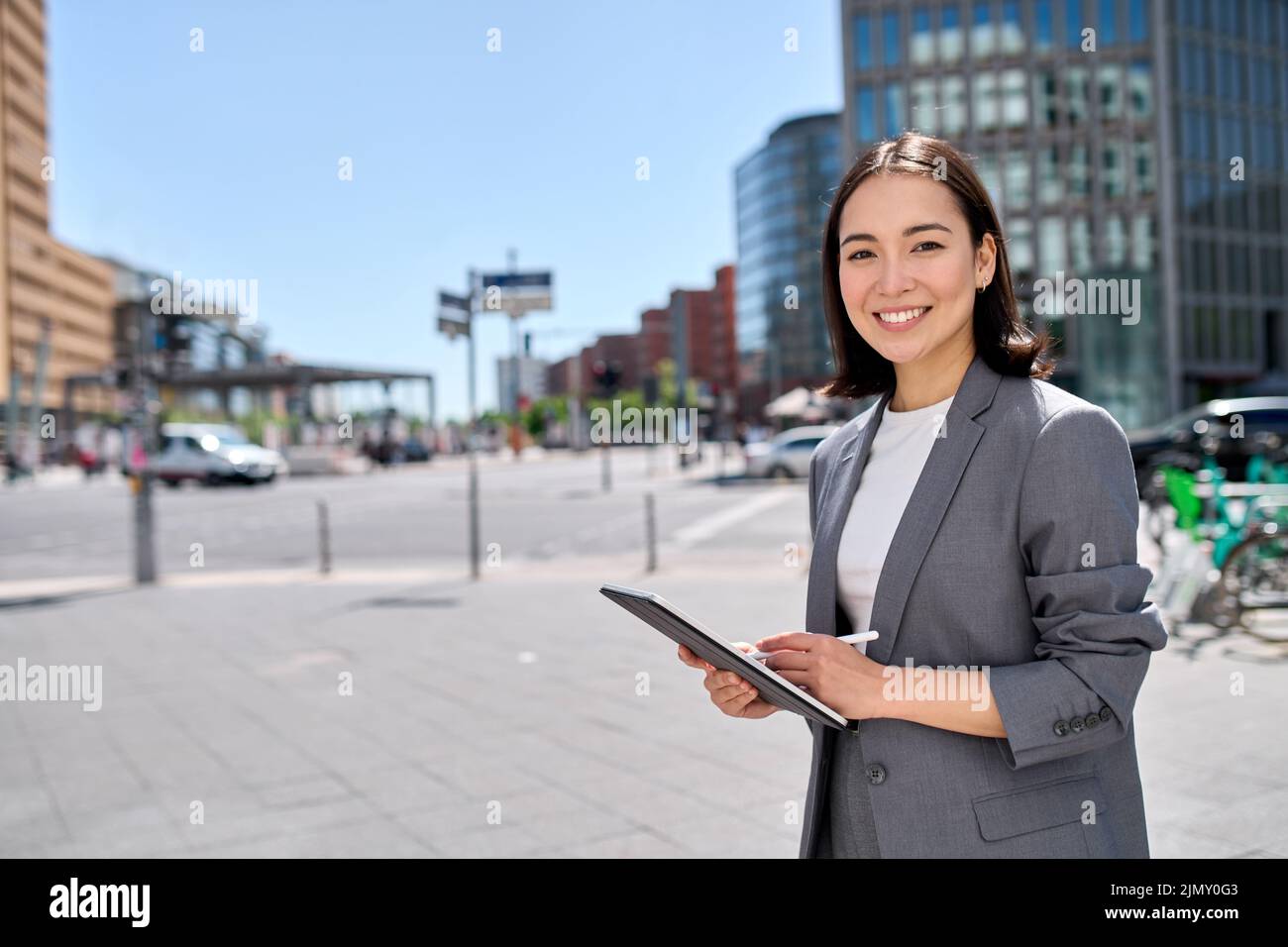 Smiling young Asian business woman standing on city street using digital tablet. Stock Photo