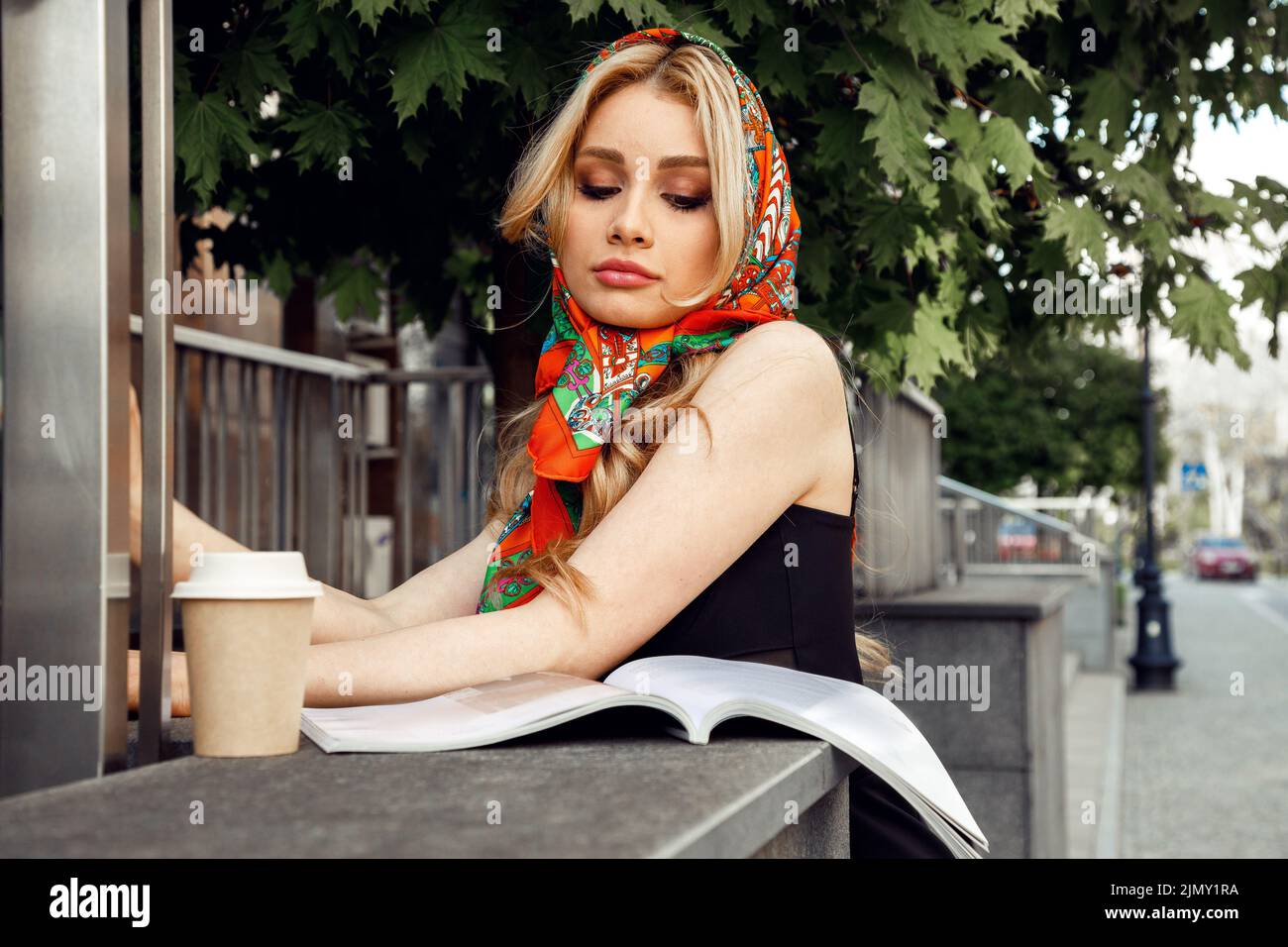 Romantic girl with bright makeup in stylish colorful headscarf looking down and reading magazine with glass of coffee in morning closeup. Paris style Stock Photo