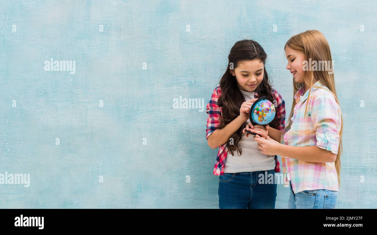 Smiling two girls standing against blue wall looking small globe Stock Photo
