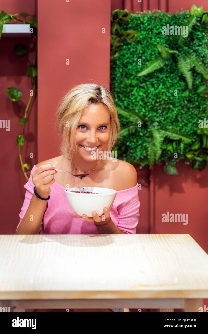 Woman smiling at camera while eating an acai smoothie bowl in a cafe. Healthy eating concept. Stock Photo