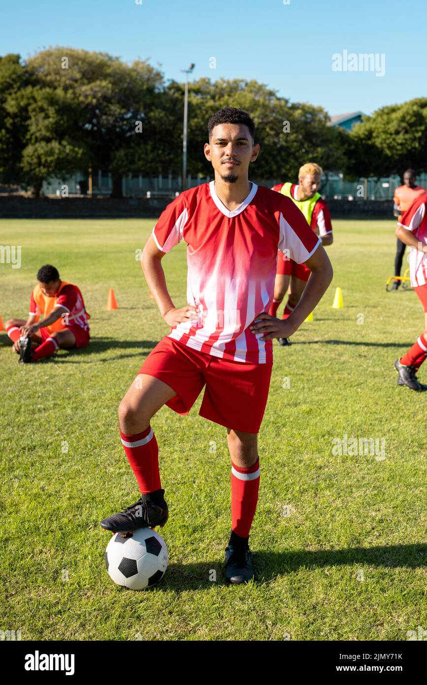Confident multiracial young player in uniform with arms akimbo and leg on soccer ball at playground Stock Photo
