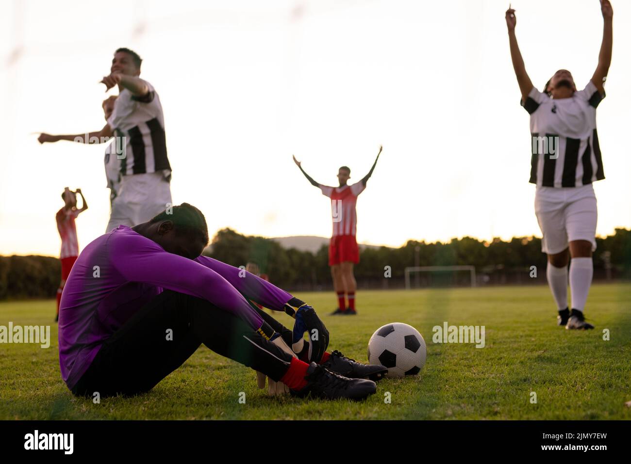 Multiracial sad goalkeeper sitting on grassy land and players with arms