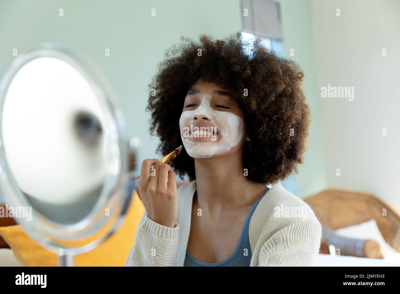 Smiling young biracial woman with afro hair applying beauty cream on face while looking at mirror Stock Photo
