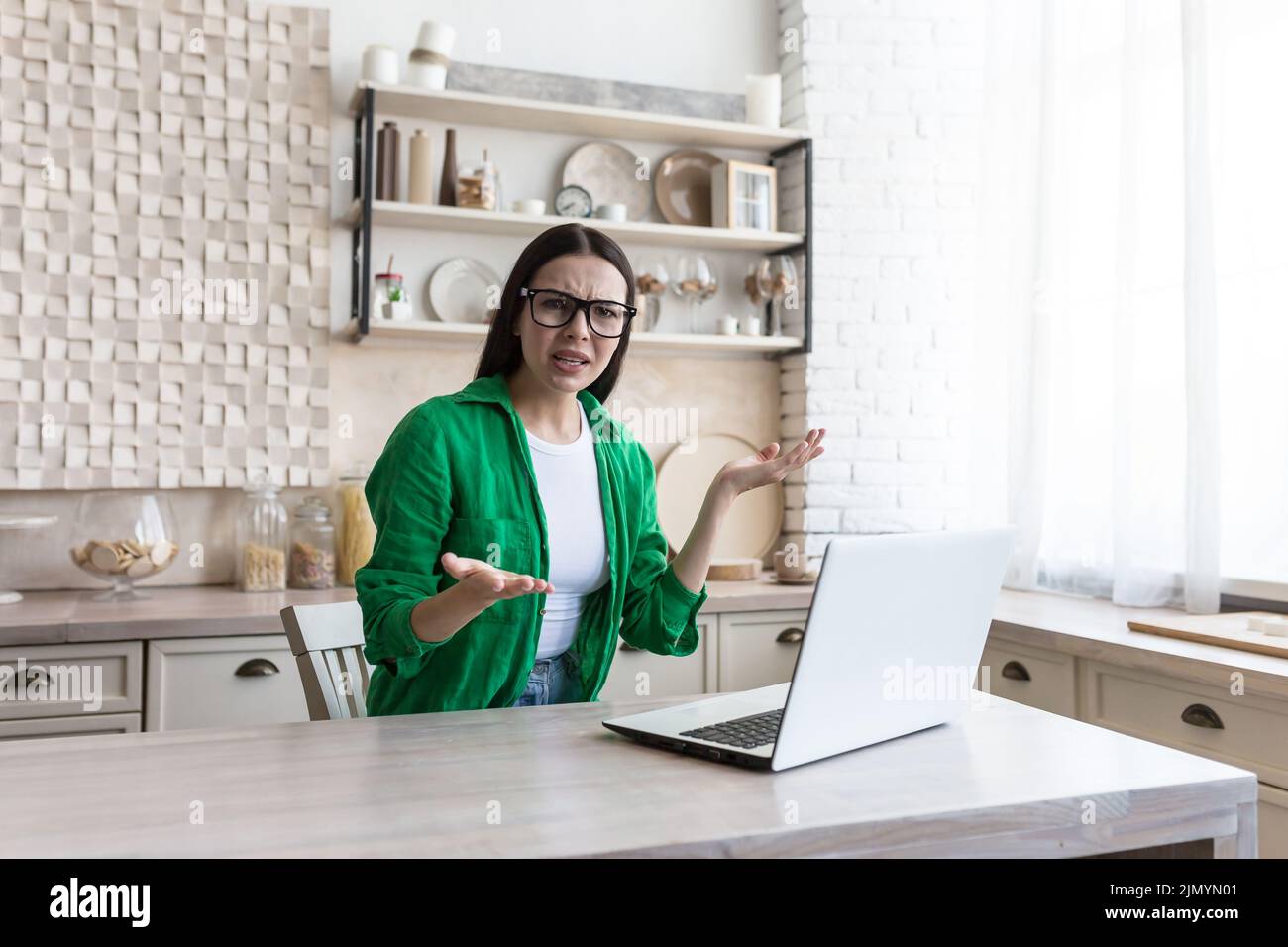 Freelance. Work at home. Confused young beautiful woman in glasses and green clothes, freelancer sitting at a table with a laptop, received bad news, upset, throws up her hands. Stock Photo