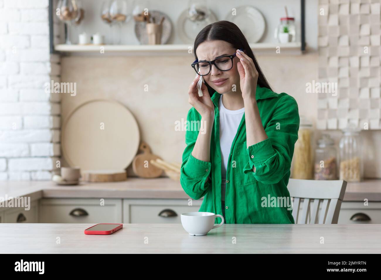 Depression, panic disorders. A young beautiful woman in glasses and a green shirt is crying at home, sitting at the kitchen table, wiping her tears with a napkin. He feels bad, feels fear and apathy Stock Photo