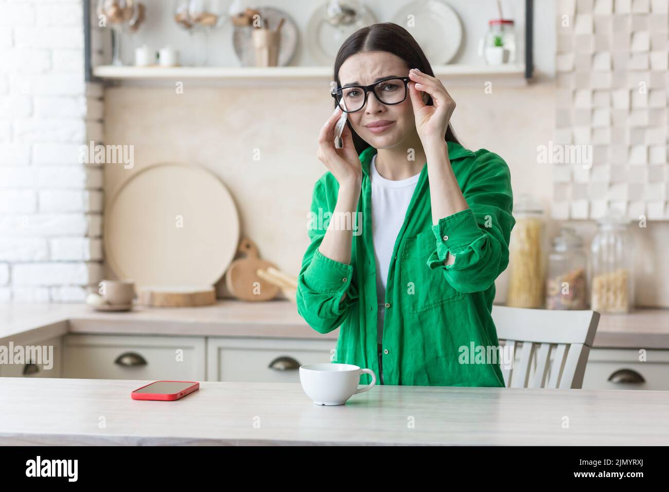 Depression, panic disorders. A young beautiful woman in glasses and a green shirt is crying at home, sitting at the kitchen table, wiping her tears with a napkin. He feels bad, feels fear and apathy Stock Photo