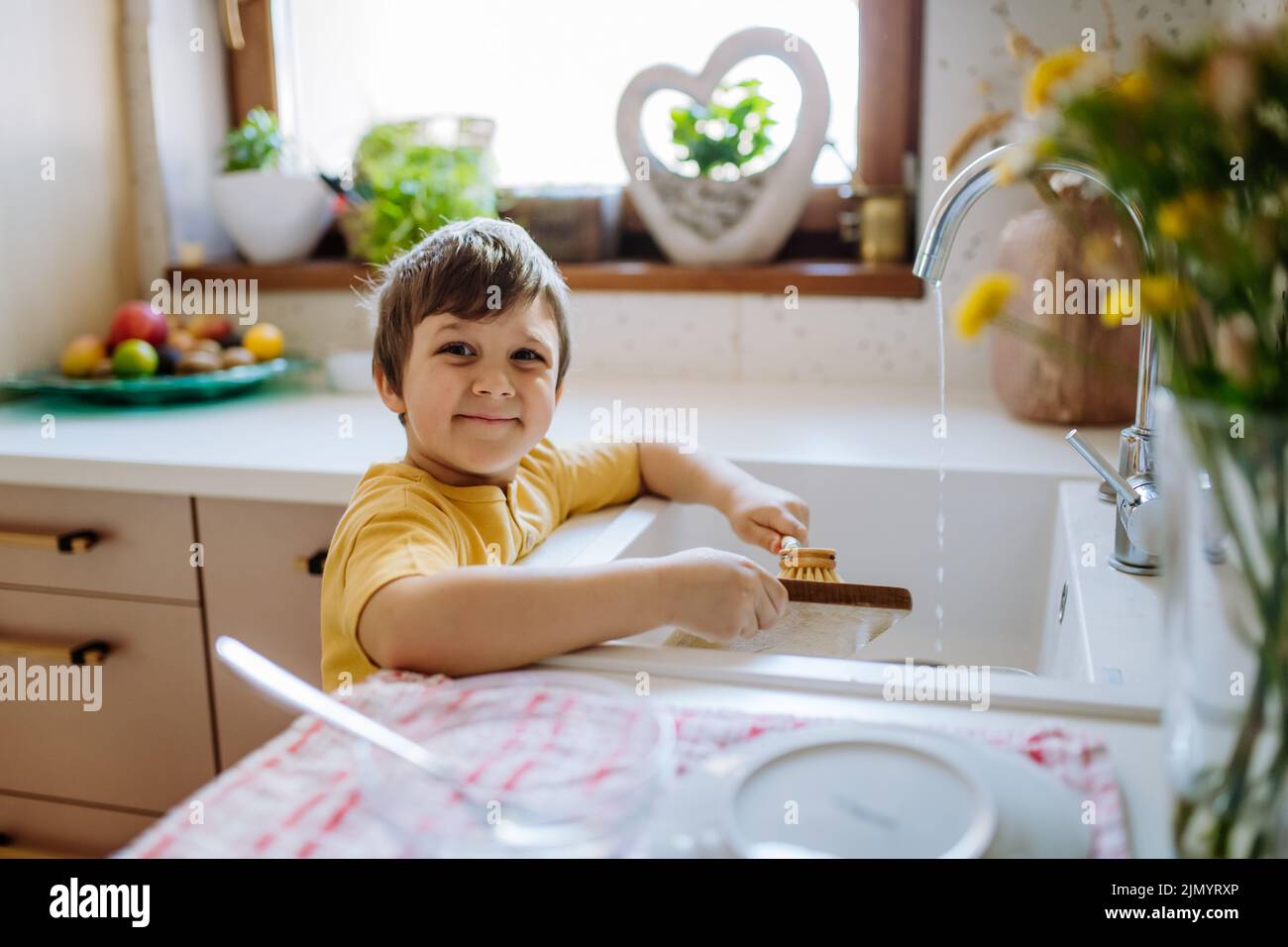 Little boy washing the dishes in sink in kitchen with wooden scrub, sustainable lifestlye. Looking at camera and smiling. Stock Photo