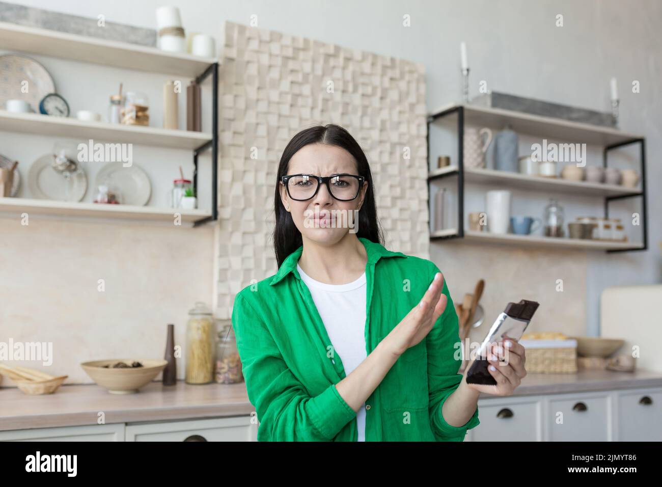 A young beautiful female nutritionist in glasses and a green shirt advises against eating chocolate. He holds a bar of chocolate and shows no with his hand. Chocolate and sweets are harmful to health Stock Photo
