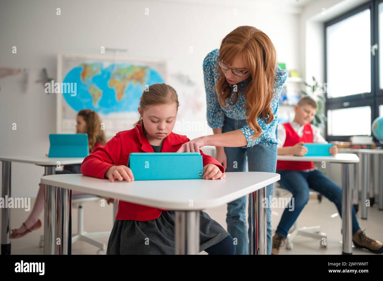 Down syndrome schoolgirl using tablet with help of teacher during class at school, integration concept. Stock Photo