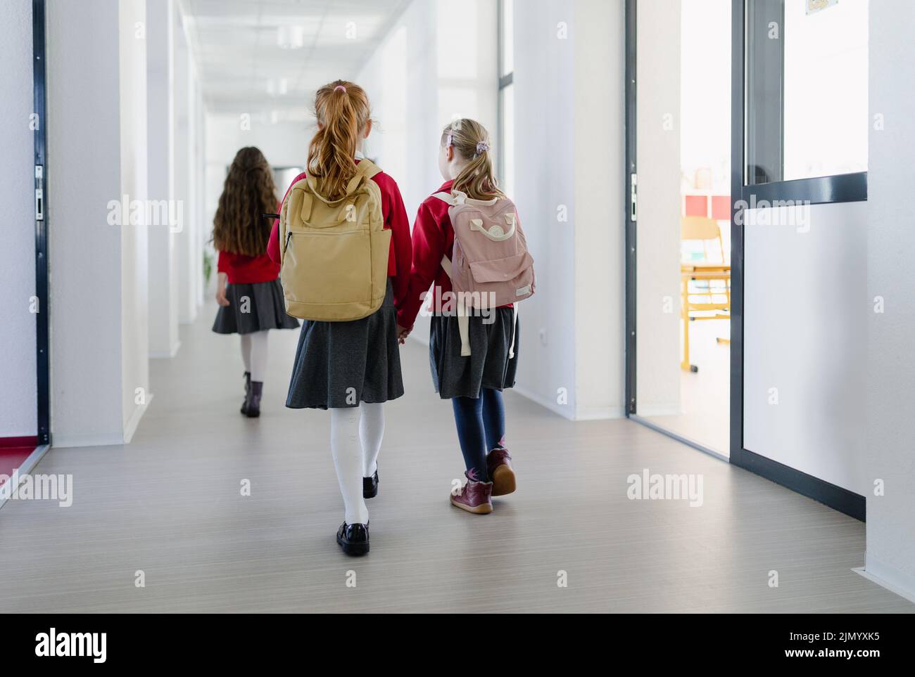 Happy schoolgirl with Down syndrome classmate in uniform walking in scool corridor with classmates, rear view. Stock Photo