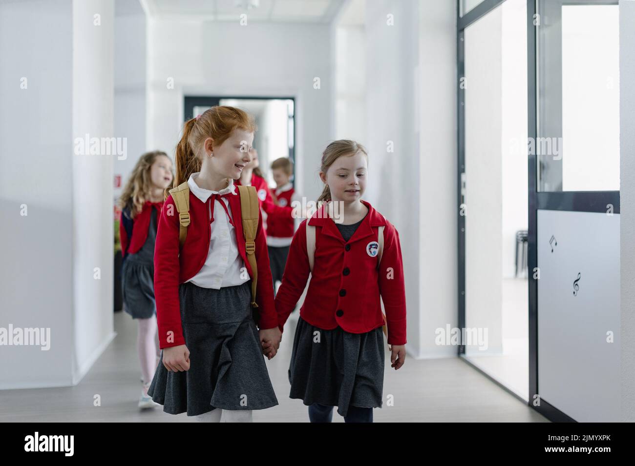 Happy schoolgrirl with Down syndrome in uniform walking in school corridor with her classmate, holding each other hands. Stock Photo