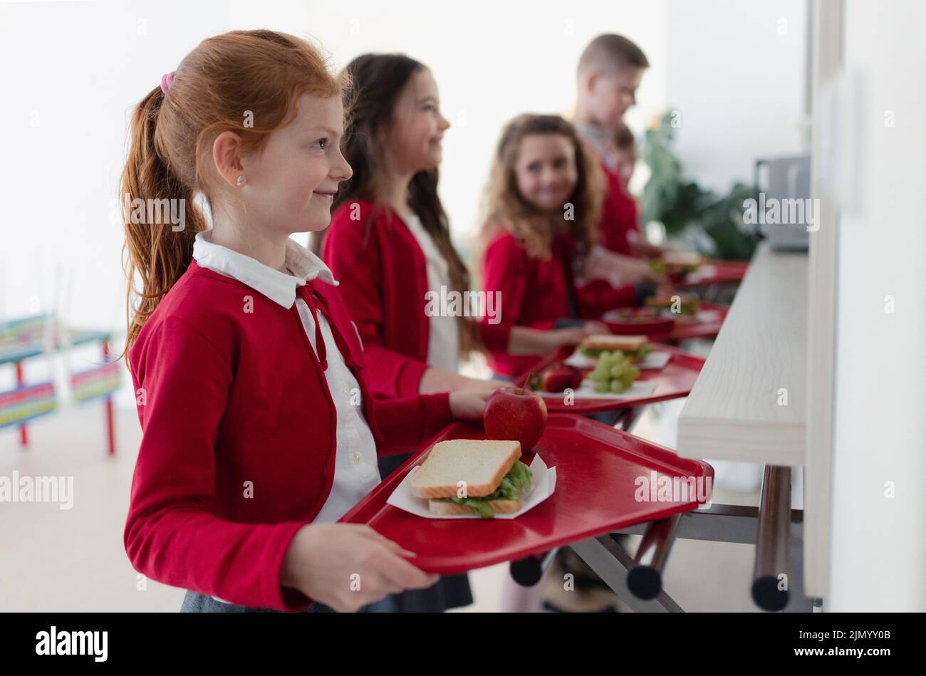 Happy schoolchildren standing in queue with trays and receiving lunch in school canteen. Stock Photo
