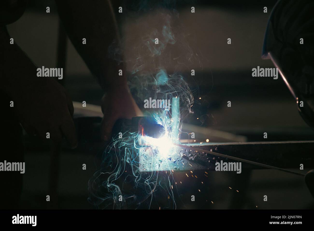 Handyman performing welding and grinding at his workplace in the workshop, while the sparks 'fly' all around him. He is wearing a protective helmet an Stock Photo