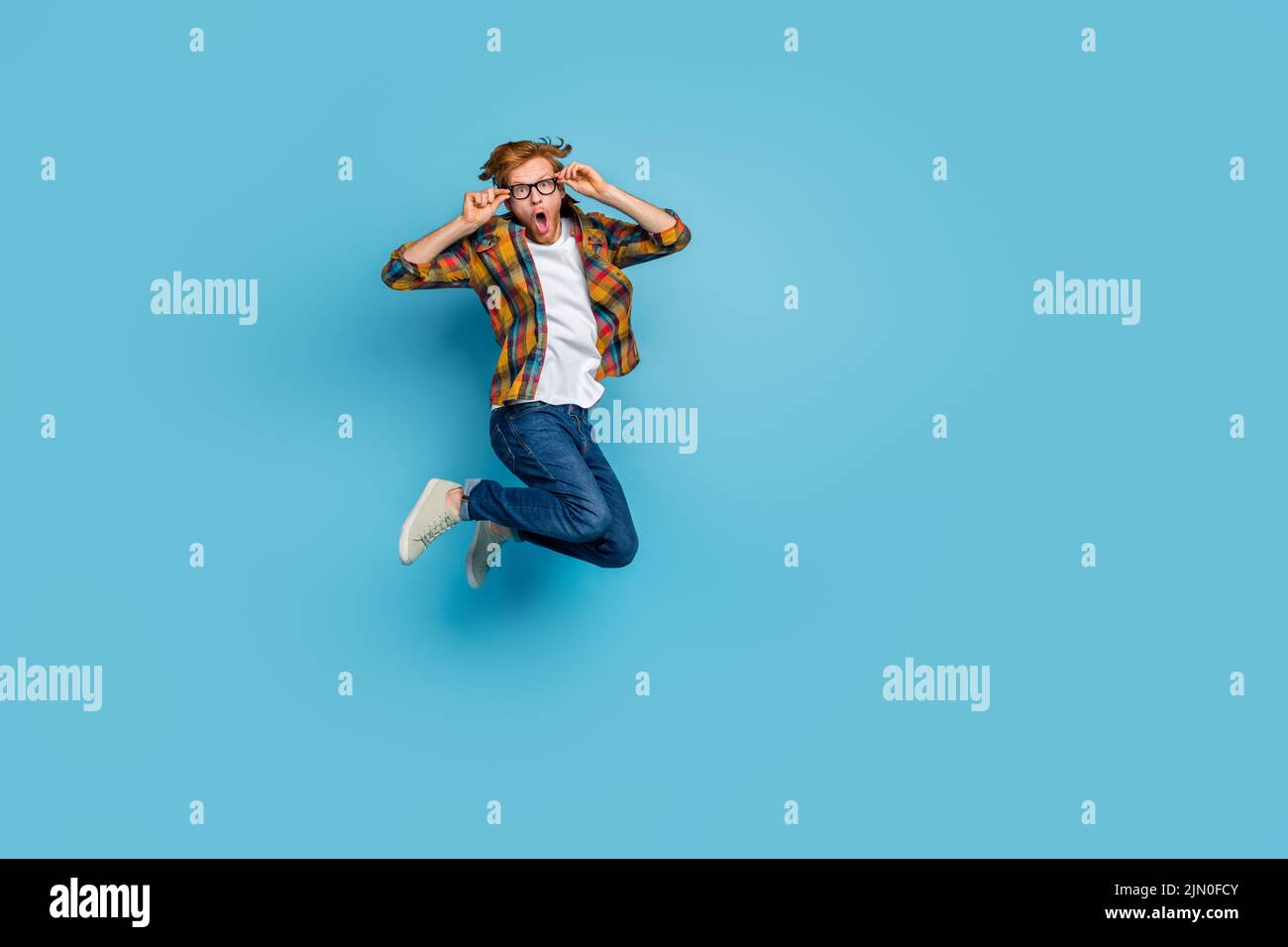 Photo of impressed excited guy with red hairstyle dressed plaid shirt holding glasses jumping shocked isolated on blue color background Stock Photo