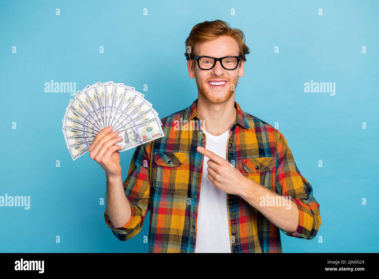 Photo of successful cheerful man with ginger hairstyle dressed plaid shirt indicating dollars in hand isolated on blue color background Stock Photo