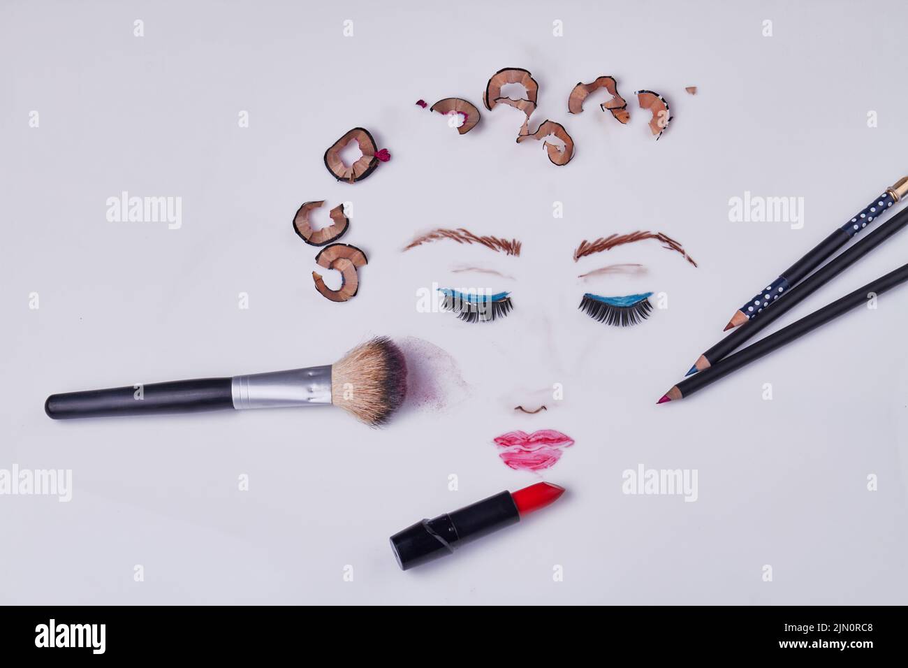 Top view makeup accessories with pencil shavings on white background. Drawn female face with her eyes closed. Stock Photo