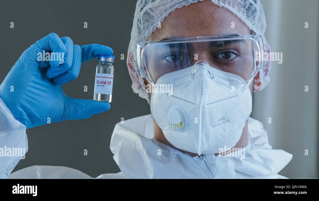 Arab man Spaniard male doctor scientist medical worker in special protective uniform sterile clothing glasses and gloves holding bottle with vaccine Stock Photo