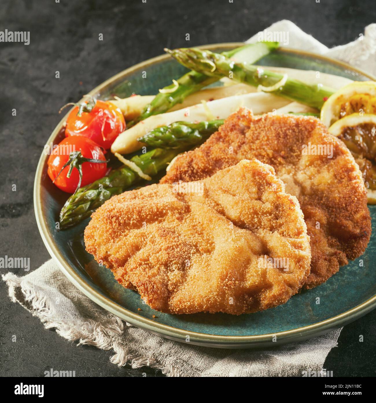 From above plate with tasty schnitzels and asparagus with cherry tomatoes and lemon served for lunch on ceramic plate on napkin against black backgrou Stock Photo