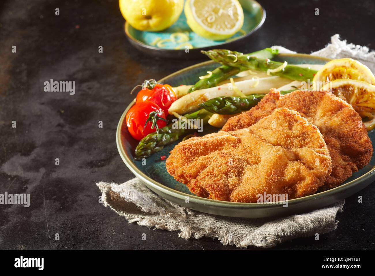 Hceramic plate of schnitzels and asparagus with cherry tomatoes and lemons placed on linen napkin on black background Stock Photo