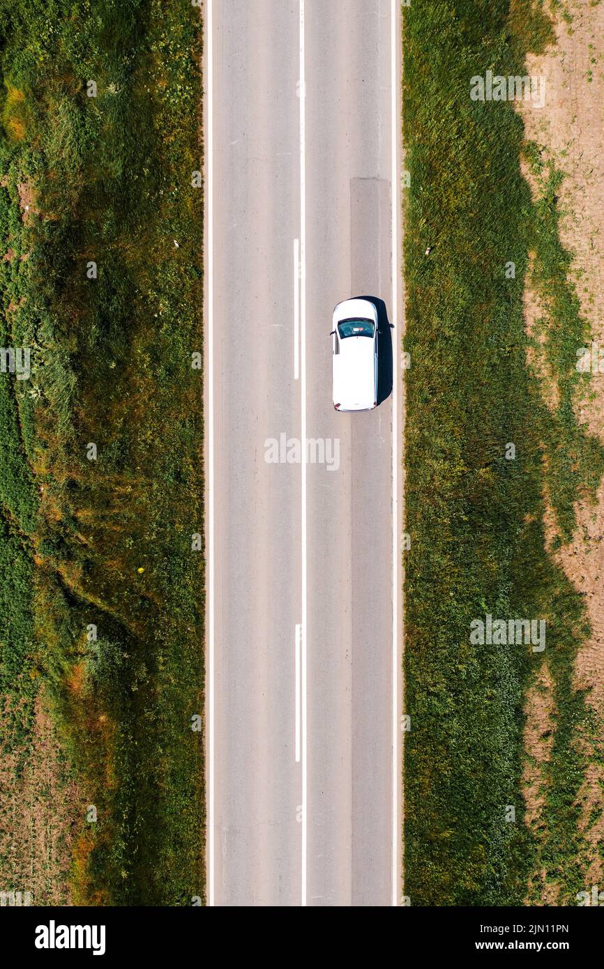 White multi-purpose vehicle is driving along the road through countryside landscape on hot summer afternoon, drone pov directly above Stock Photo