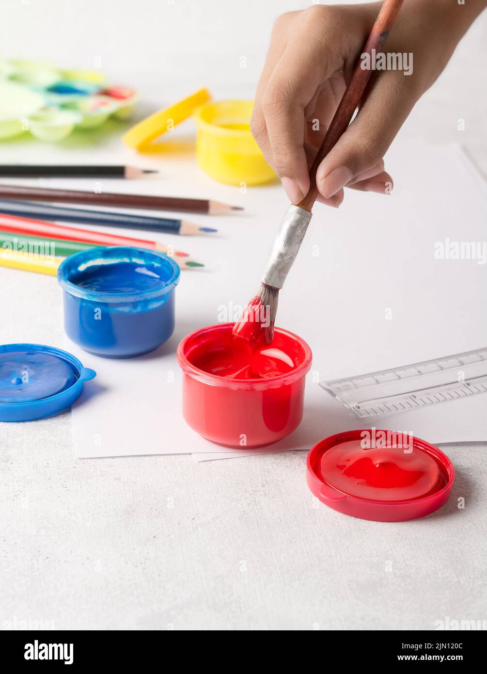 close-up of young child hand dip paint brush into acrylic paint and gouache color jars, on white background, soft-focus with copy space, kids hobbies Stock Photo