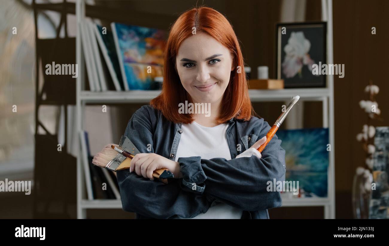 Portrait Caucasian girl confident talented artist woman painter with red hair holding equipment for painting paintbrush trowel spatula crossing arms Stock Photo