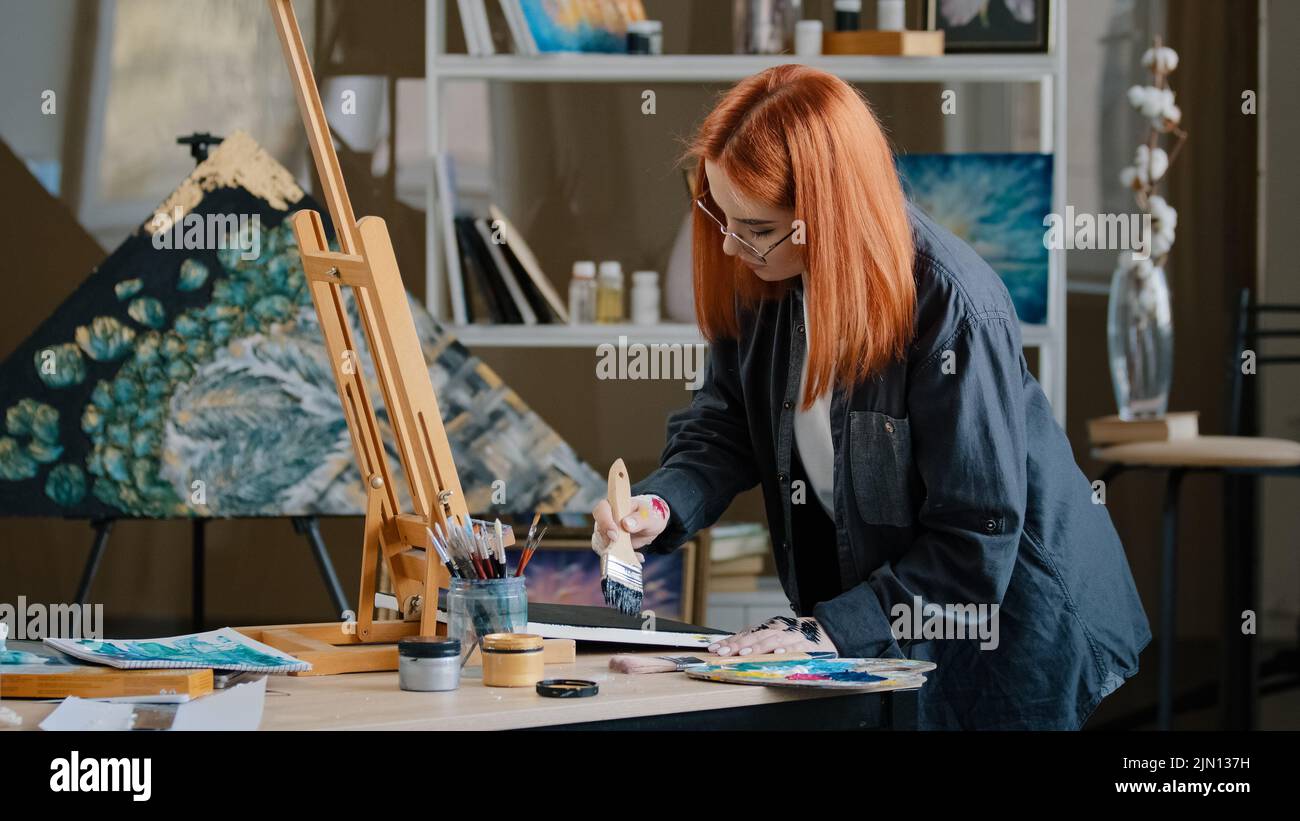 Inspired busy focused redhead girl artist woman painter in art studio background paints with wide brush on canvas with acrylic oil colorful paint puts Stock Photo