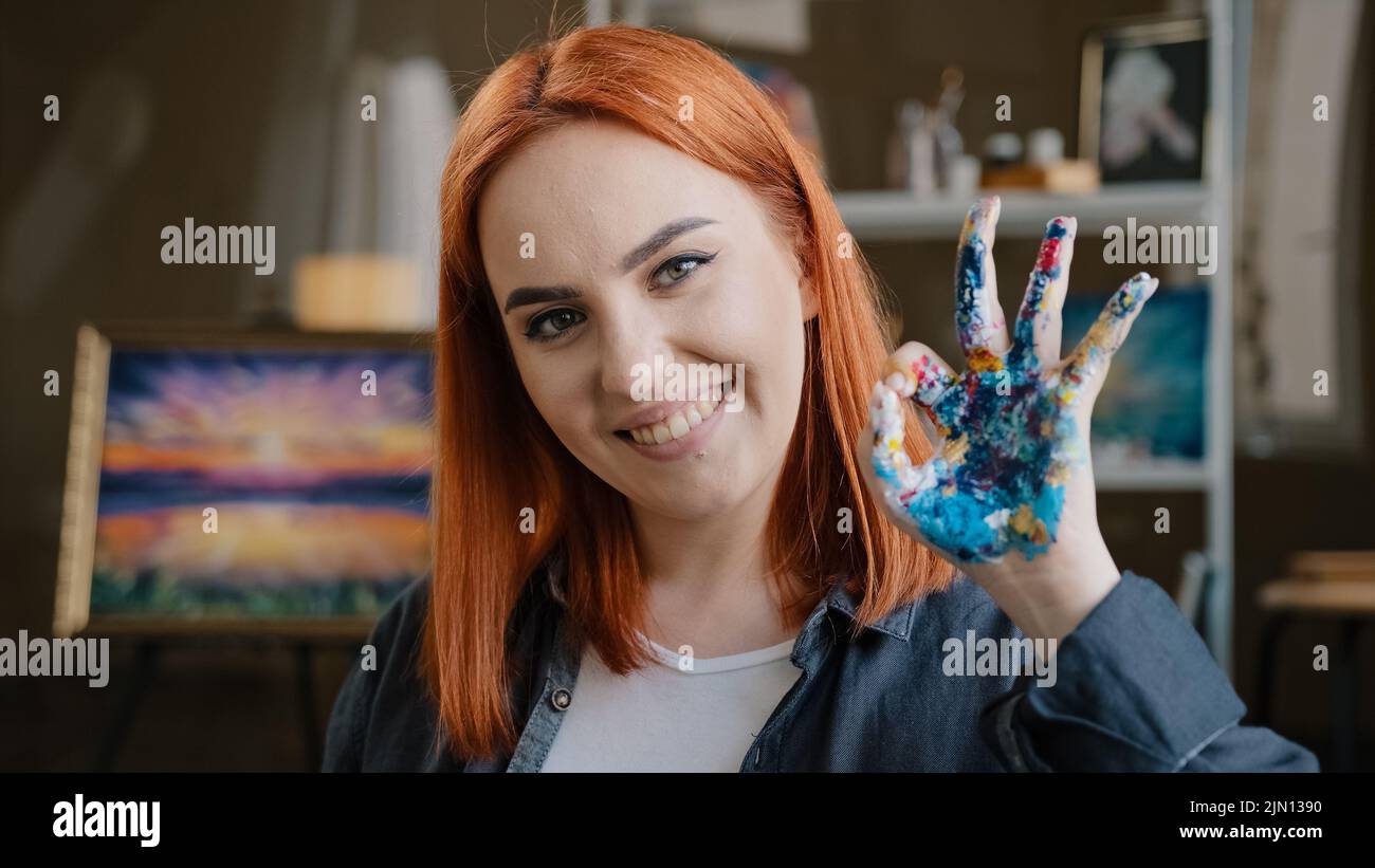 Red-haired girl talented artist female painter stands posing in art studio workshop looking at camera show okay gesture dirty palm in acrylic oil Stock Photo
