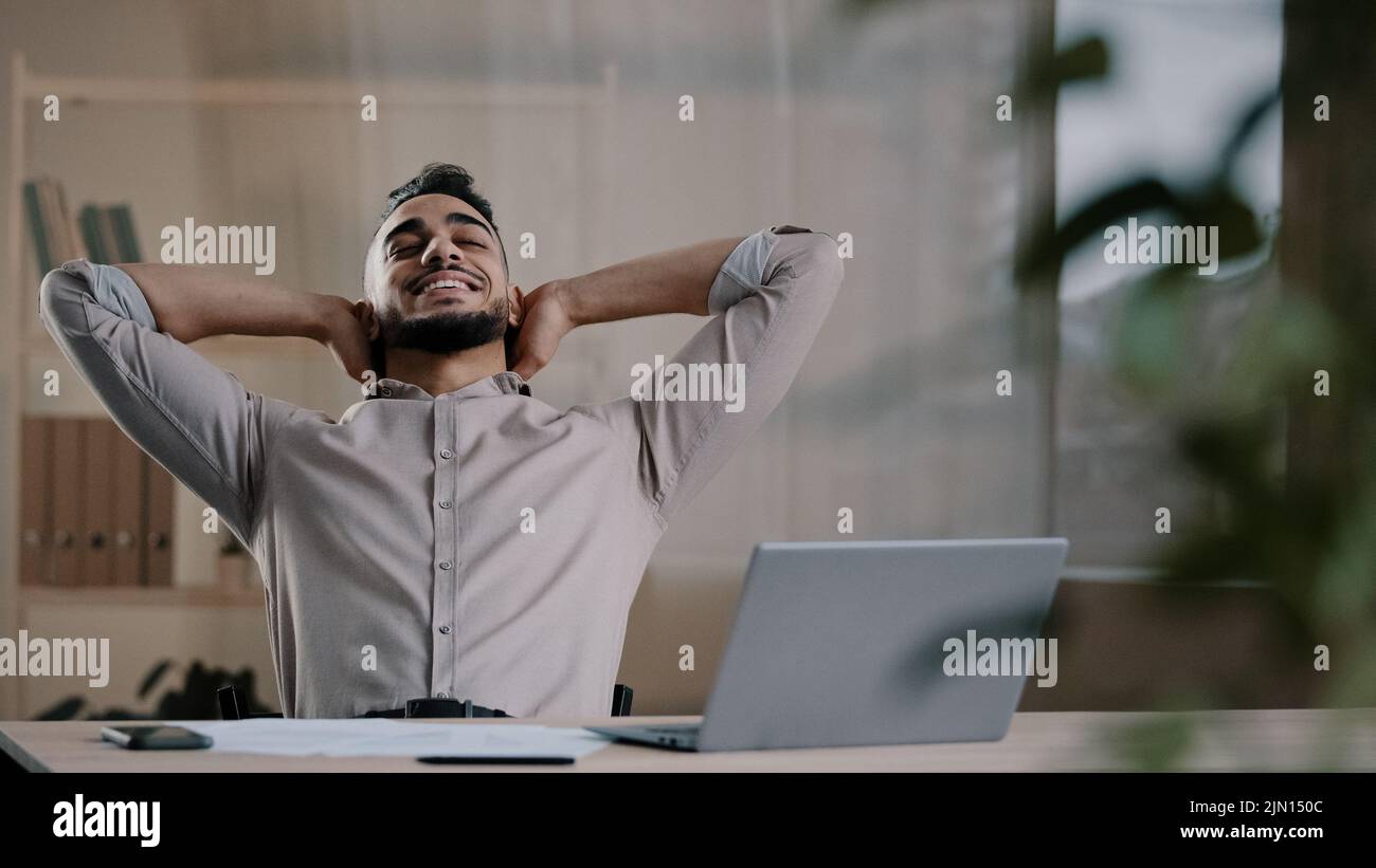 Happy satisfied arab man worker businessman hispanic guy finished work task on computer sit at home office put hands behind head lean on chair take Stock Photo