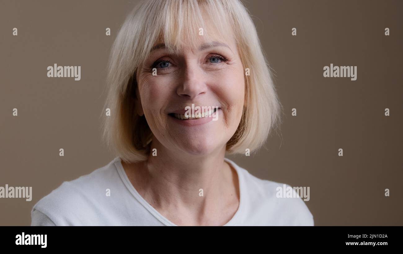 Happy mature middle-aged blonde smiling caucasian 60s woman looking at camera posing in studio portrait, cheerful older grey-haired female granny in Stock Photo