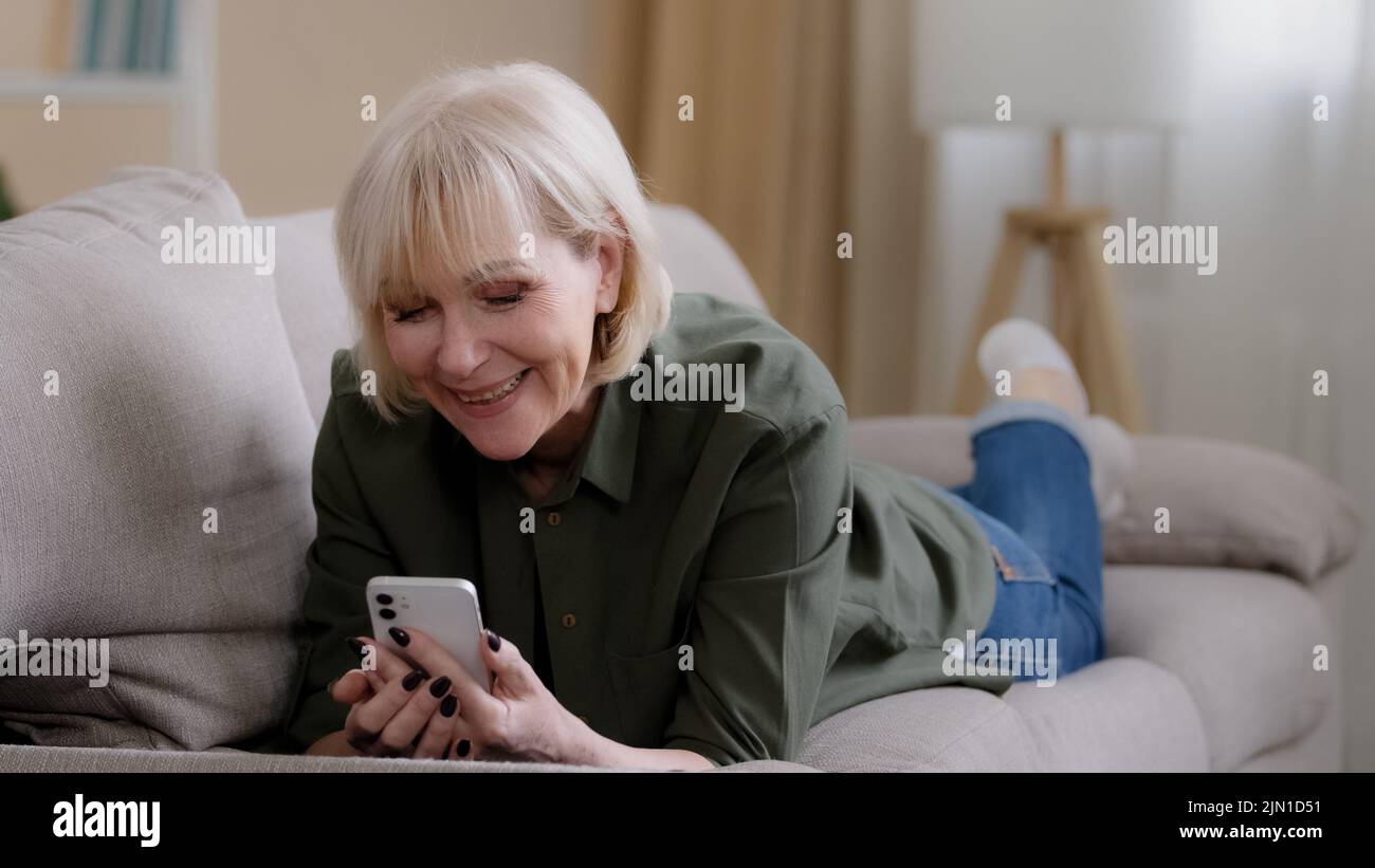 Old happy Caucasian grandmother 60 years senior woman 50s lady blonde elderly female lies on couch at home enjoys remote mobile conversation video Stock Photo