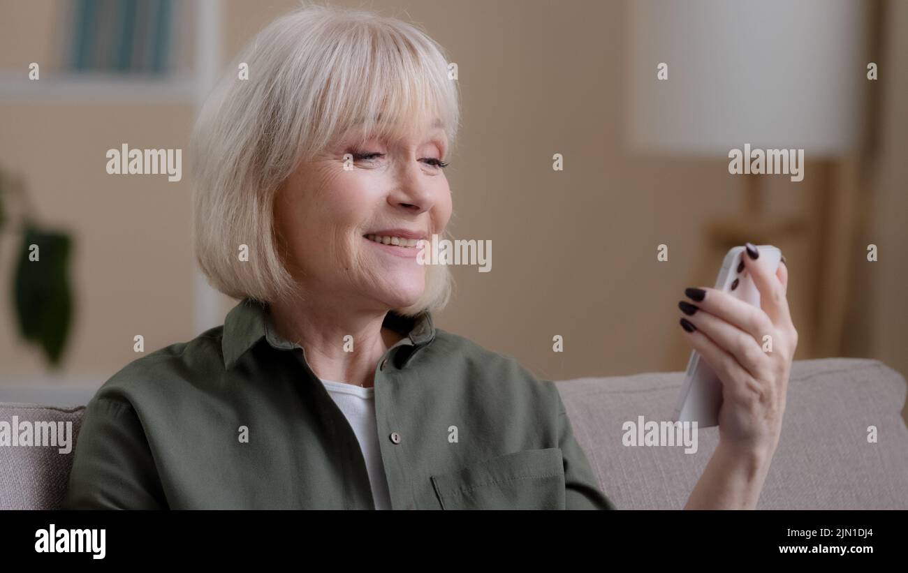 Old talking Caucasian blonde wrinkled grandmother mature senior woman elderly female has video call with mobile phone app at home remote conference Stock Photo