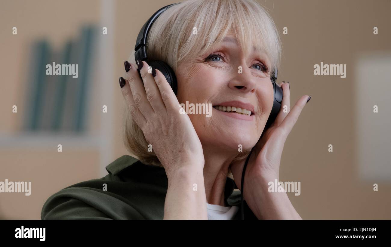 Portrait happy relaxed blonde 60s grandmother dancing old Caucasian woman in headphones carefree senior lady in earphones listening to music enjoys Stock Photo