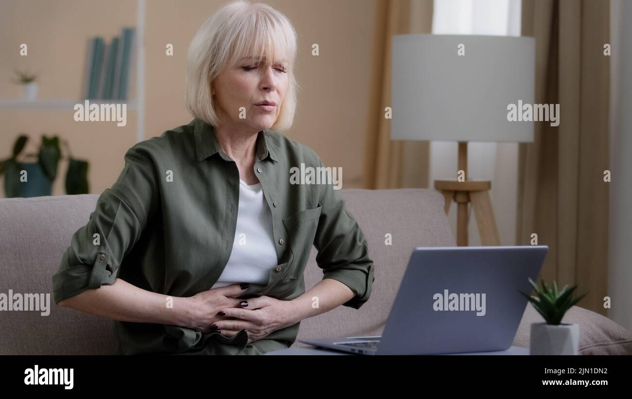 Sick mature woman with laptop bad pain hold belly stomach suffer with hurt abdomen ache at home, old lady feels gastritis pancreatitis symptom sit on Stock Photo