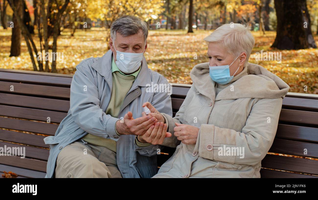 Elderly couple in medical masks in autumn park spouses disinfect hands with antiseptic sitting on bench retirees care about health personal hygiene Stock Photo