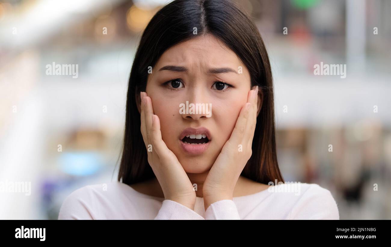 Headshot female emotional face asian ethnic korean girl 20s woman model frightened lady scared fear horror disgust holding hands behind head from Stock Photo