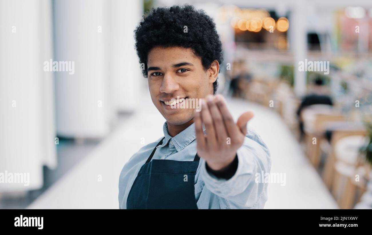 Friendly african american young guy male waiter salesman restaurant cafe kitchen bar worker in apron looking at camera showing gesture her you come Stock Photo