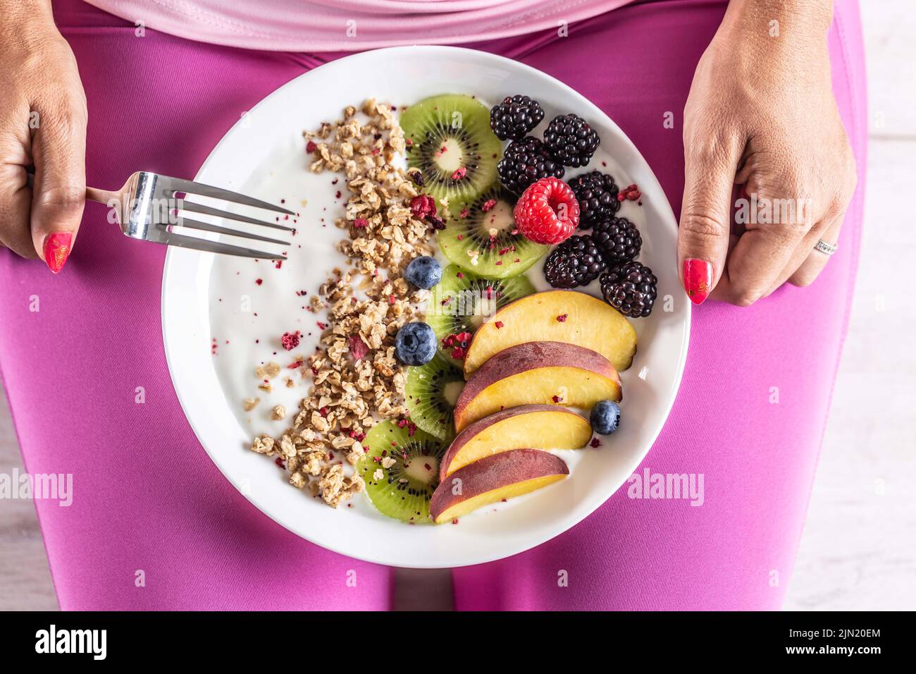 A woman has a healthy breakfast after morning exercise. Yogurt, blackberry muesli, raspberries, blueberries, kiwi and peaches in a white bowl. Stock Photo