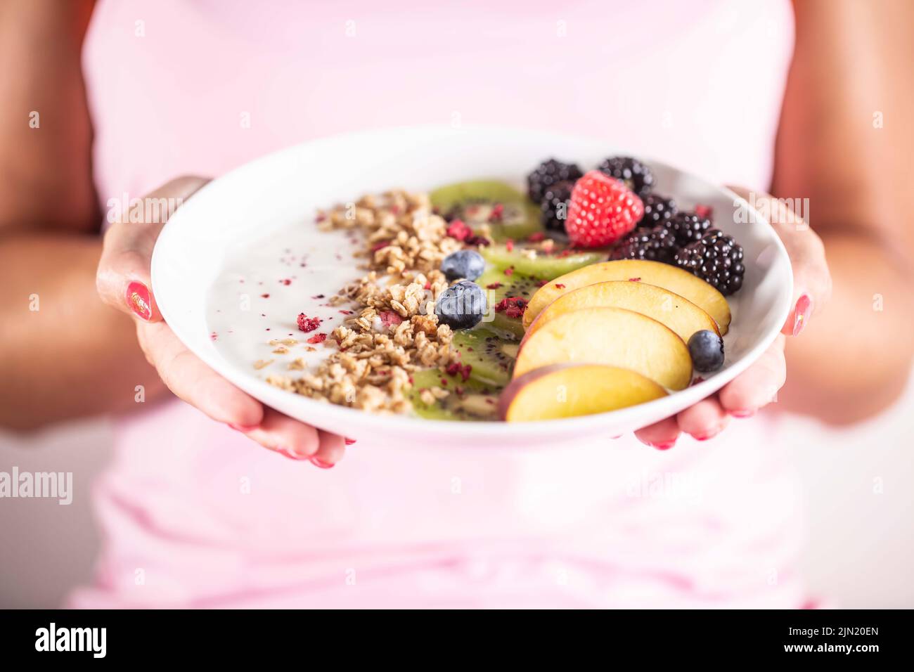 Yogurt, blackberry muesli, raspberries, blueberries, kiwi and peaches in a white bowl holding young woman. Stock Photo