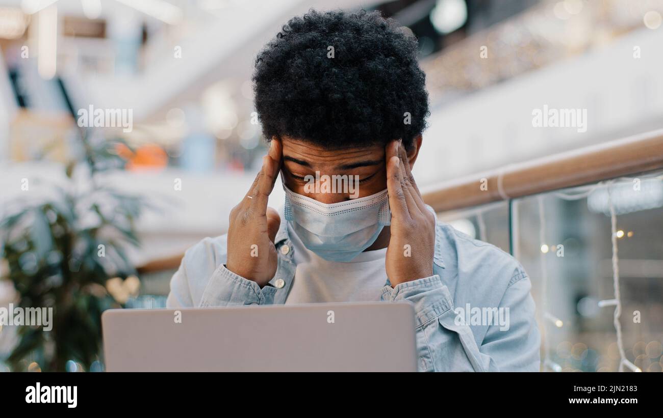 African american sad business man guy student wearing medical mask working with laptop in corporate office holding head feeling headache suffering Stock Photo