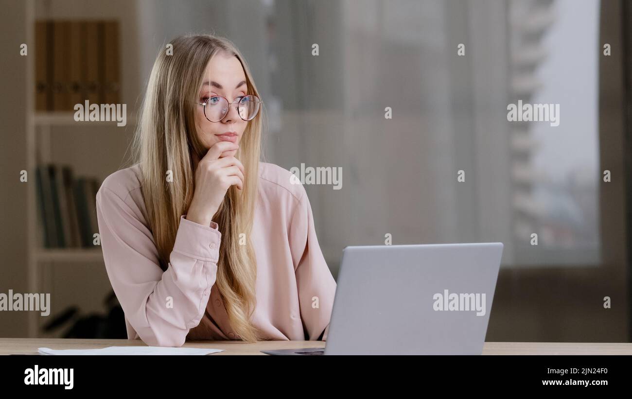 Caucasian millennial business woman female student girl writer journalist wearing glasses sitting at table at office home with laptop thinks idea Stock Photo