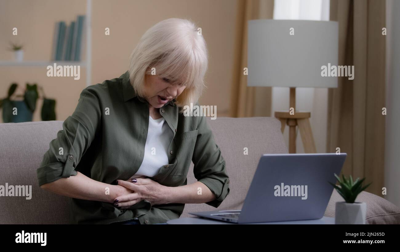 Sick 50s woman with laptop bad pain hold belly stomach suffer with hurt abdomen ache at home, old lady feels gastritis pancreatitis symptom sit on Stock Photo