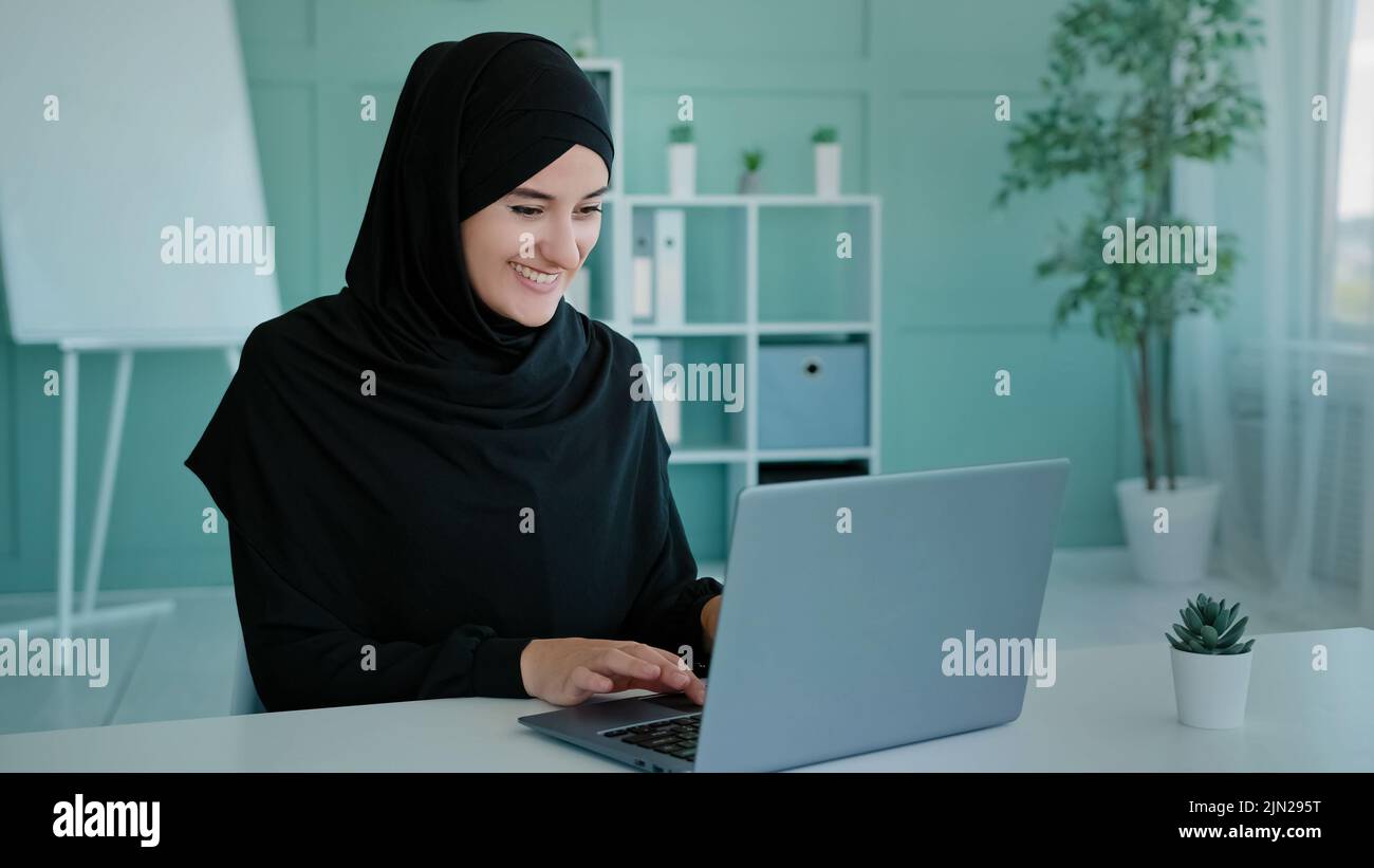 Islamic Muslim successful woman in black hijab celebrate victory triumph look at laptop screen shocked close mouth with astonish winning lottery bid Stock Photo