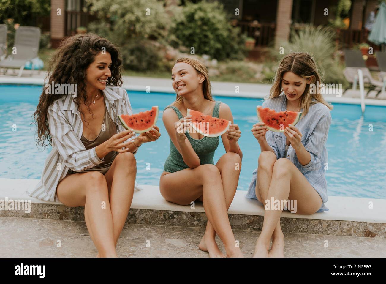Three cute young women sitting on by the swimming pool and eating watermelon in the house backyard Stock Photo