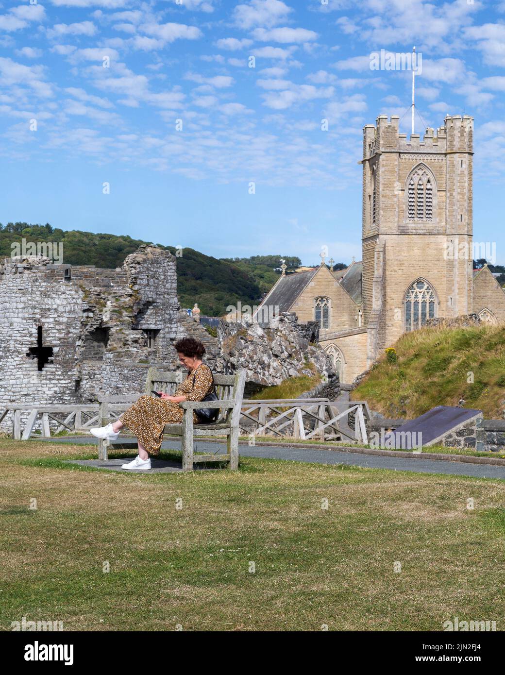 A woman looks at her phone while sitting on a bench in the grounds of Aberystwyth castle. St. Michael's church is seen beyond part of the castle ruin. Stock Photo