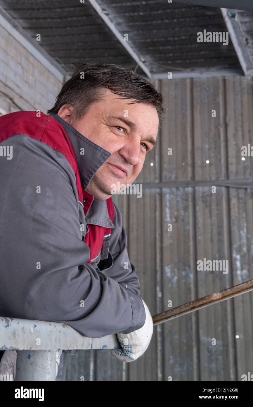 A tired middle-aged male worker in a protective dirty uniform looks into the distance while working in a warehouse, factory, vertical frame. Portrait Stock Photo