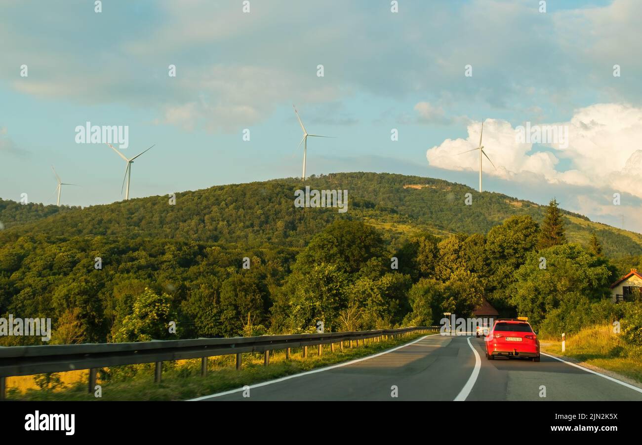 Red car drives on road against green hillside with windmill farm. Producing ecological electrical energy among green landscape under blue sky Stock Photo